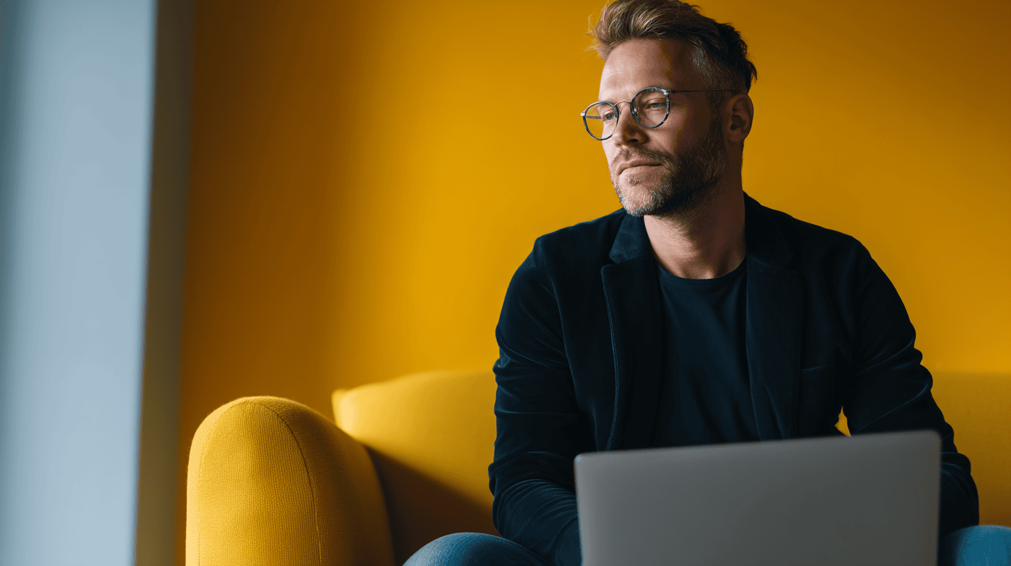 Man wearing glasses and a dark blazer sitting on a yellow chair, looking thoughtfully out of frame, with a laptop in front of him and a vibrant yellow wall in the background.