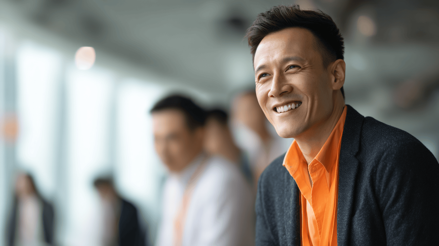 Smiling man in an orange shirt and dark blazer, looking to the side in a bright, modern office environment with blurred colleagues in the background.