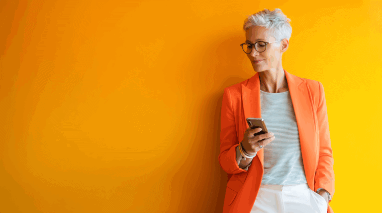 Confident woman with short gray hair and glasses, wearing an orange blazer and holding a smartphone while standing against a bright yellow wall