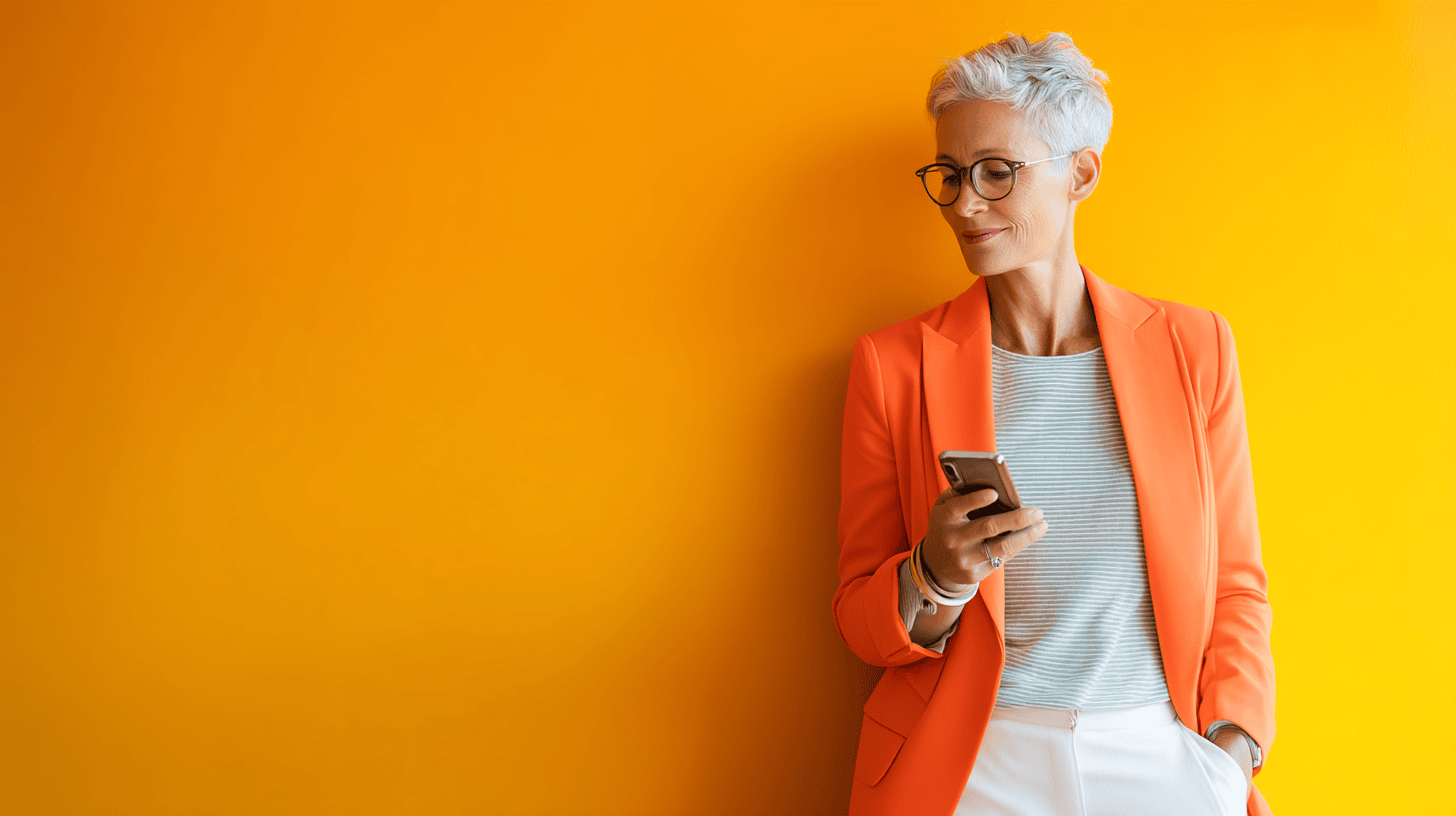 Confident woman with short gray hair and glasses, wearing an orange blazer and holding a smartphone while standing against a bright yellow wall