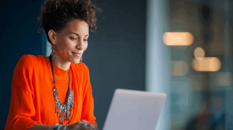 a woman in an orange shirt and necklace looking at a laptop