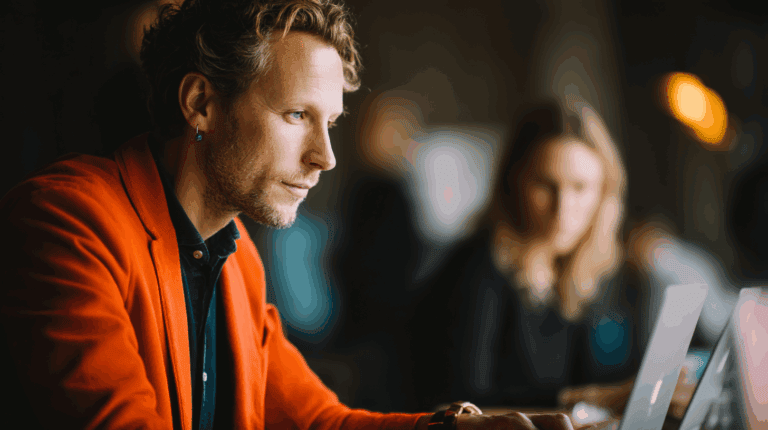 A man with curly hair is focused on his work while wearing an orange blazer. He sits at a desk with laptops nearby, with a woman blurred in the background. The setting appears to be a modern workspace, capturing a professional atmosphere.