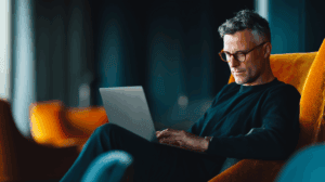 A casually dressed man with short, wavy hair and glasses is sitting comfortably in a vibrant orange chair, focused on a laptop in front of him. The background is softly blurred, featuring additional seating and curtains, creating a modern, relaxed atmosphere.