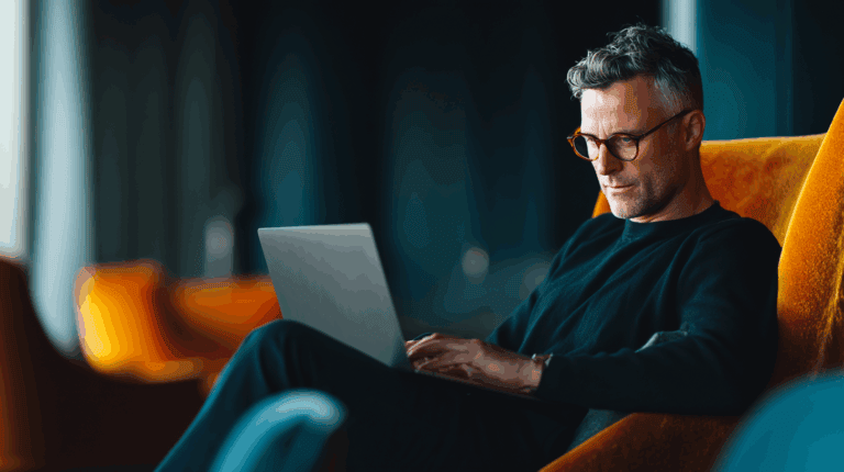 A casually dressed man with short, wavy hair and glasses is sitting comfortably in a vibrant orange chair, focused on a laptop in front of him. The background is softly blurred, featuring additional seating and curtains, creating a modern, relaxed atmosphere.