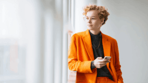 A fashionable person with curly hair wearing stylish glasses stands by a window, holding a smartphone. They are dressed in an orange blazer over a dark gray sweater, exuding a modern and confident vibe. Soft natural light highlights their thoughtful expression against a neutral background.