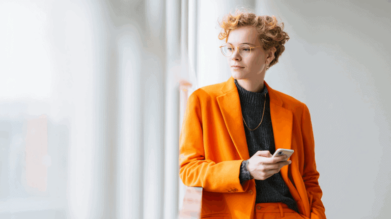 A fashionable person with curly hair wearing stylish glasses stands by a window, holding a smartphone. They are dressed in an orange blazer over a dark gray sweater, exuding a modern and confident vibe. Soft natural light highlights their thoughtful expression against a neutral background.