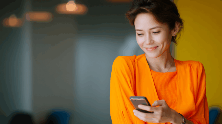 A smiling woman wearing an orange top holds a smartphone while looking at it. She is in a well-lit indoor space with a blurred background, enhancing the cheerful atmosphere.
