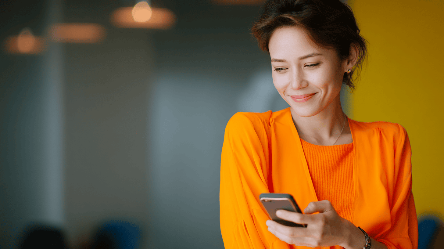 A smiling woman wearing an orange top holds a smartphone while looking at it. She is in a well-lit indoor space with a blurred background, enhancing the cheerful atmosphere.