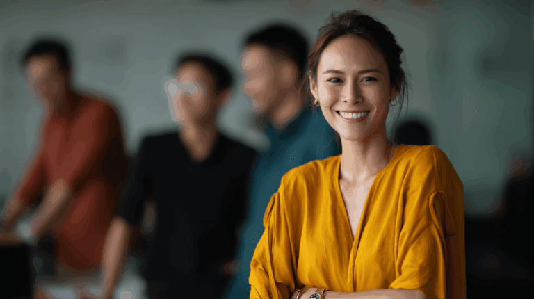 A smiling woman wearing a vibrant yellow blouse stands confidently in an office setting. She has her arms crossed and is surrounded by blurred figures of colleagues in the background, suggesting a collaborative environment. The overall atmosphere feels warm and inviting, reflecting teamwork and positivity.