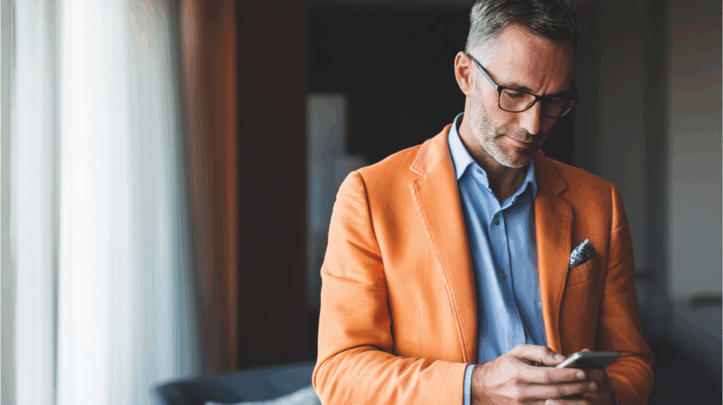A well-dressed man with short gray hair, wearing glasses, is focused on his smartphone. He is dressed in an orange blazer over a light blue shirt, with a patterned pocket square. Soft natural light filters through sheer curtains in the background, creating an elegant atmosphere.