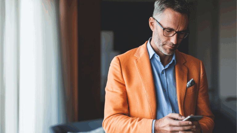A well-dressed man with short gray hair, wearing glasses, is focused on his smartphone. He is dressed in an orange blazer over a light blue shirt, with a patterned pocket square. Soft natural light filters through sheer curtains in the background, creating an elegant atmosphere.