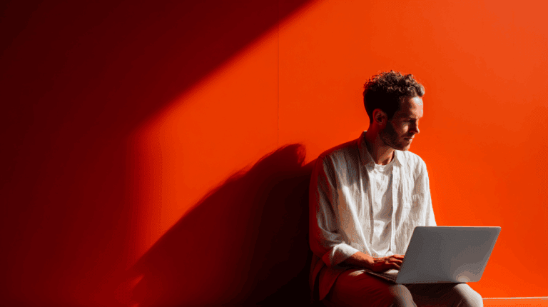 A young man sits against a vibrant orange wall, using a laptop. He is dressed in a light-colored, loose-fitting shirt and has short, curly hair. Sunlight creates a dramatic shadow, highlighting his focused expression as he works or studies. The scene conveys a calm and creative atmosphere.