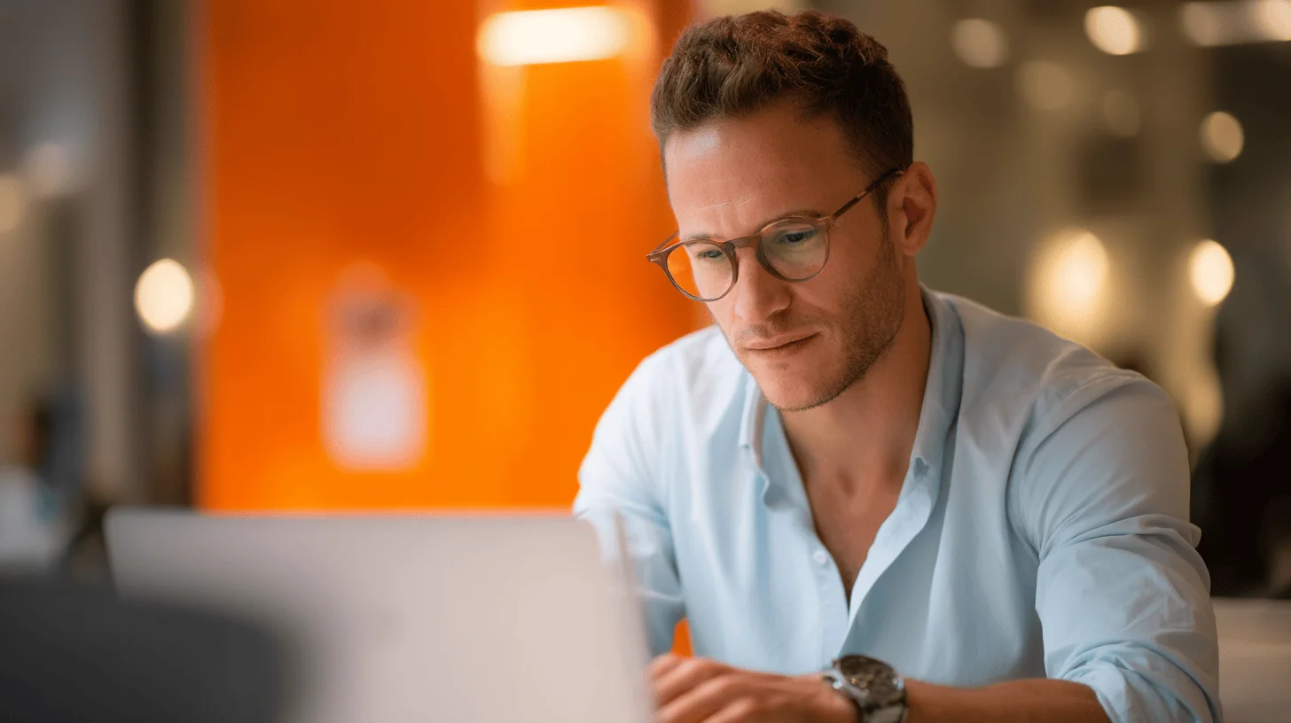 A young man with glasses, wearing a light blue shirt, is focused on his laptop screen in a softly lit indoor environment. An orange wall is in the background, creating a warm ambiance. The man appears engaged in his work, indicating concentration or analysis.