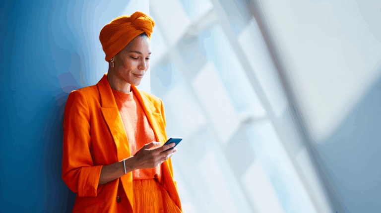 A woman stands against a blue wall, wearing a vibrant orange suit and matching headwrap. She is holding a smartphone and looking at the screen, smiling softly. Bright light filters in through large windows behind her, creating a warm and inviting atmosphere.