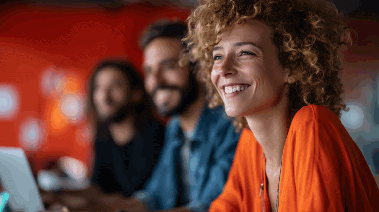 A close-up of a smiling woman with curly hair, wearing an orange top, sitting at a table with two blurred colleagues in the background. They are working on laptops, and the backdrop features a vibrant red wall, creating a lively and collaborative atmosphere.