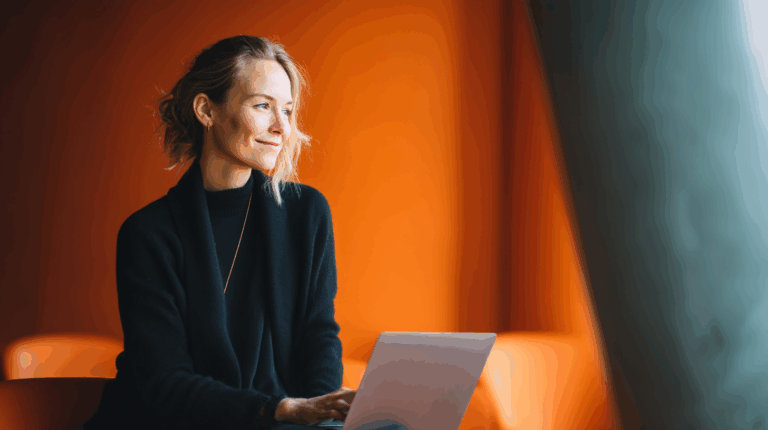 A smiling woman with wavy hair sits comfortably while working on a laptop. She is dressed in a black sweater, and the background features warm orange hues, enhancing a cozy atmosphere. Natural light highlights her thoughtful expression as she engages in her work.