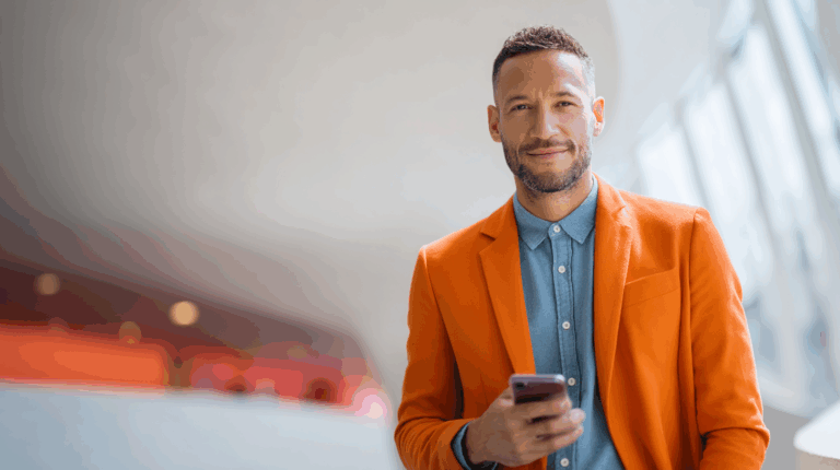 A man with a well-groomed beard stands confidently, wearing an orange blazer over a light blue shirt. He is holding a smartphone and smiling slightly. The background features a modern, softly lit interior with abstract shapes, creating a vibrant and professional atmosphere.