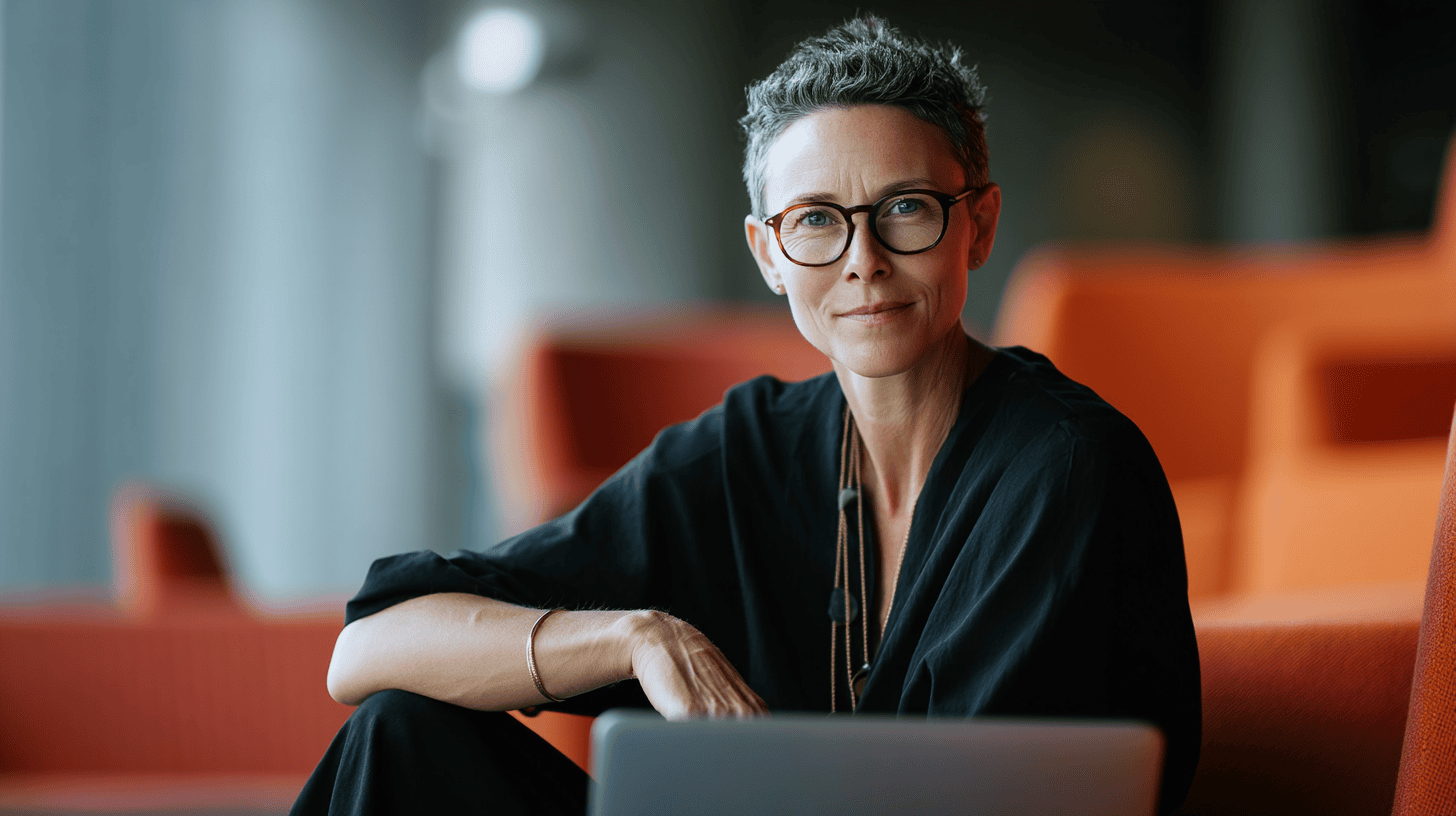 A confident woman with short, curly hair is seated at a table, wearing black attire. She has round glasses and is smiling softly at the camera. A laptop is in front of her, and the background features vibrant orange seating and a modern interior space, creating a warm, inviting atmosphere.