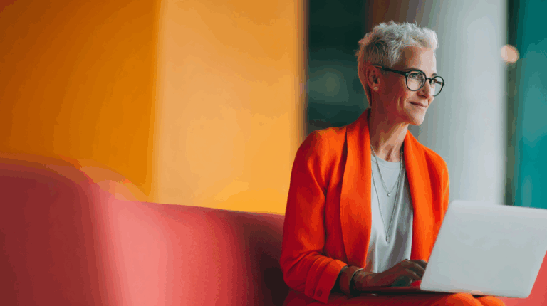A smiling woman with short, silver hair and glasses sits on a pink couch, working on a laptop. She wears a bright orange blazer over a light-colored top. The background features warm, colorful tones, creating a modern and inviting workspace atmosphere.