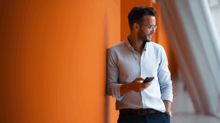 A well-dressed man with glasses leans against an orange wall, casually looking at his smartphone. His light blue shirt contrasts with the vibrant background, and he appears relaxed and engaged in his device. Natural light highlights the modern architectural setting.