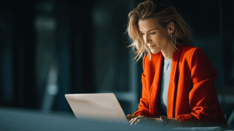 A woman with shoulder-length blonde hair, wearing an orange blazer over a gray top, is focused on her laptop in a modern office setting. Soft lighting highlights her features, conveying a sense of concentration and professionalism. The background is slightly blurred, emphasizing her engagement with the screen.