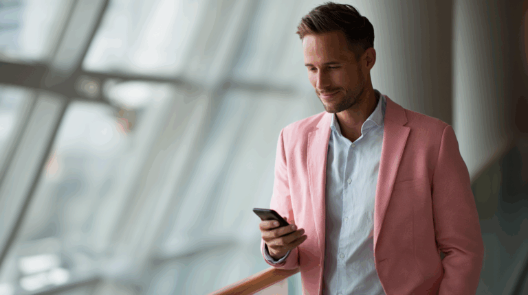 A well-dressed man in a pink blazer and light blue shirt is standing next to a large window, looking at his smartphone with a slight smile. The background features blurred glass panels, suggesting a modern interior space.
