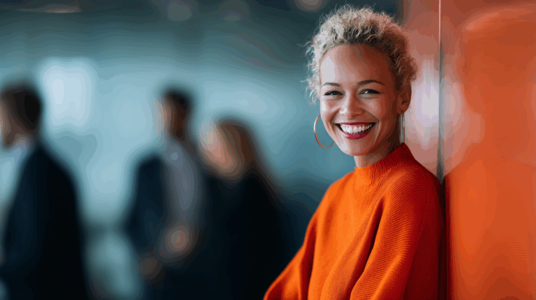 A cheerful woman with curly hair wearing an orange sweater leans against a wall, smiling brightly. The background features blurred figures in a professional setting, creating a lively atmosphere. The image conveys positivity and confidence.