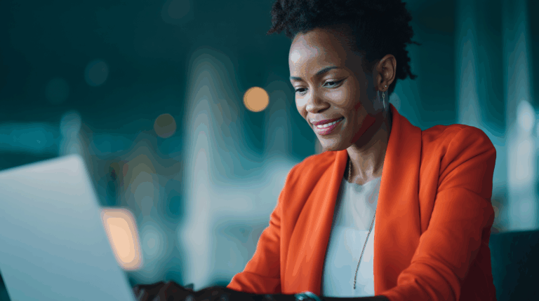 A woman wearing an orange blazer smiles while using a laptop in a modern office setting. The background features soft lighting and a blurred view of the workspace, creating a professional and positive atmosphere.