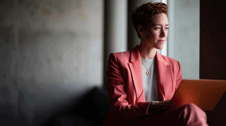 A professional person wearing a pink suit sits by a window, focused on a laptop. The background is softly blurred, highlighting their thoughtful expression. Natural light illuminates the scene, creating a warm and inviting atmosphere.