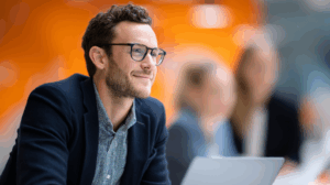 A smiling man with curly hair and glasses sits attentively in a modern office setting. He wears a dark blazer over a patterned shirt and is focused on his laptop. In the background, two blurred colleagues can be seen, suggesting a collaborative work environment with an orange backdrop.