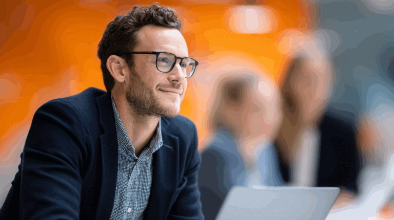 A smiling man with curly hair and glasses sits attentively in a modern office setting. He wears a dark blazer over a patterned shirt and is focused on his laptop. In the background, two blurred colleagues can be seen, suggesting a collaborative work environment with an orange backdrop.