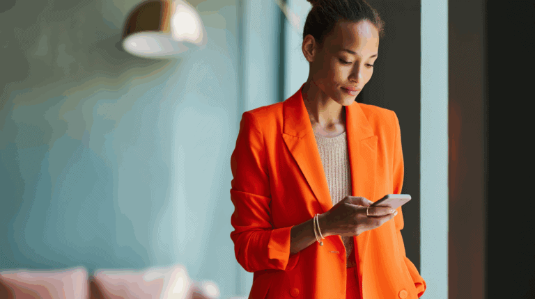 A woman in an orange blazer stands by a window, looking at her smartphone. She has a neutral expression and wears a light beige top underneath her blazer. The background features a blurred soft blue wall and a hint of pink seating. The lighting creates a warm, inviting atmosphere.