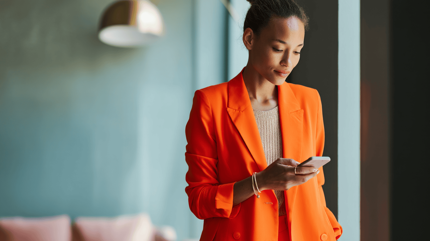 A woman in an orange blazer stands by a window, looking at her smartphone. She has a neutral expression and wears a light beige top underneath her blazer. The background features a blurred soft blue wall and a hint of pink seating. The lighting creates a warm, inviting atmosphere.