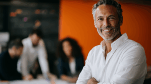 A smiling middle-aged man with salt-and-pepper hair and a beard stands confidently with his arms crossed. He is wearing a white collared shirt. In the background, three blurred individuals are collaborating, with an orange wall behind them. The setting suggests a modern, creative workspace.