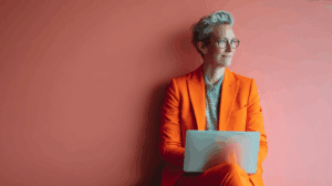 A person wearing an orange suit and glasses is sitting against a coral-colored wall, looking thoughtfully at a laptop placed on their lap. They have short, styled gray hair and are dressed in a patterned shirt beneath the suit. The scene conveys a sense of professionalism and contemplation.