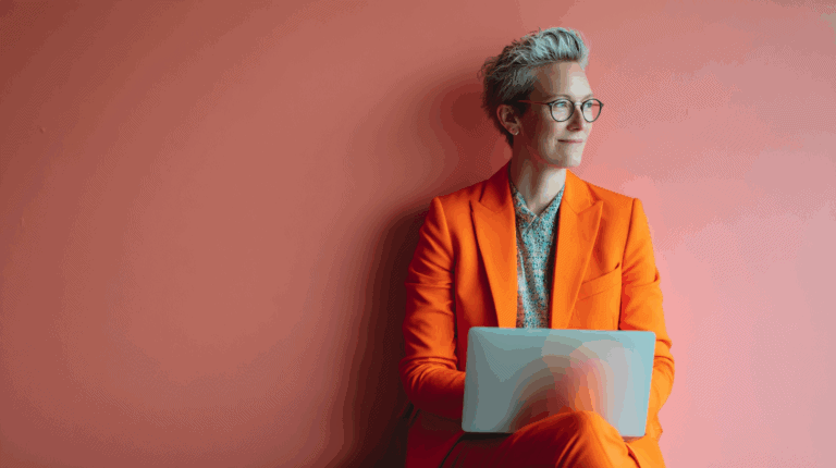 A person wearing an orange suit and glasses is sitting against a coral-colored wall, looking thoughtfully at a laptop placed on their lap. They have short, styled gray hair and are dressed in a patterned shirt beneath the suit. The scene conveys a sense of professionalism and contemplation.