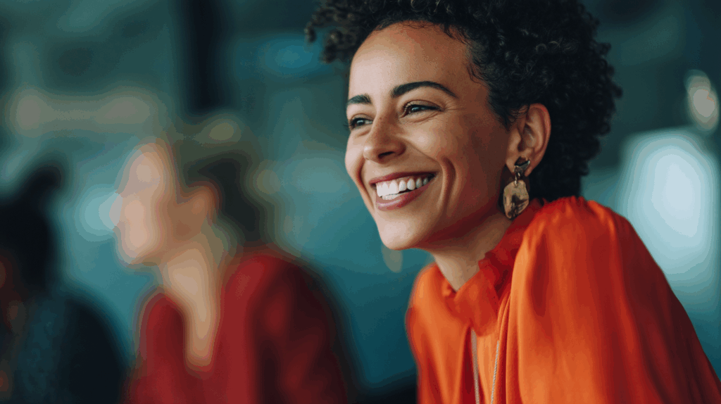 A woman with curly hair smiles brightly while wearing an orange blouse and statement earrings. She is seated at a table, with a blurred background that includes other people. The atmosphere appears warm and friendly, suggesting a moment of joy or engagement in a social setting.