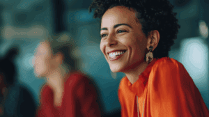 A woman with curly hair smiles brightly while wearing an orange blouse and statement earrings. She is seated at a table, with a blurred background that includes other people. The atmosphere appears warm and friendly, suggesting a moment of joy or engagement in a social setting.
