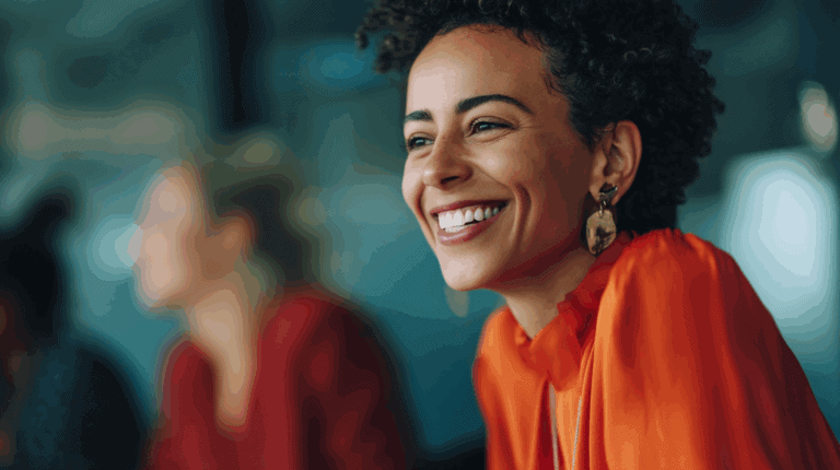 A woman with curly hair smiles brightly while wearing an orange blouse and statement earrings. She is seated at a table, with a blurred background that includes other people. The atmosphere appears warm and friendly, suggesting a moment of joy or engagement in a social setting.