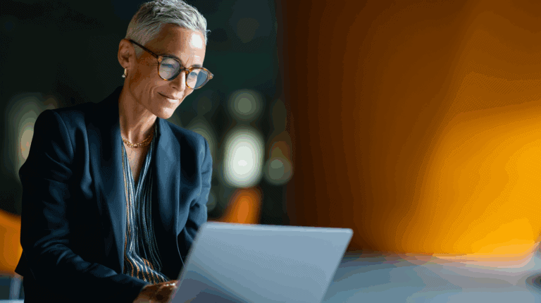 A professional-looking woman with short, silver hair is seated at a table, focused on her laptop. She wears round glasses and a stylish black blazer over a patterned top. The background features warm, muted tones, creating a modern, sophisticated atmosphere.