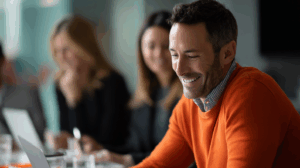 A smiling man in an orange sweater sits at a conference table, working on a laptop. In the background, two blurred colleagues are visible, one with long hair and the other in business attire, creating an atmosphere of collaboration and focus in a modern office setting.