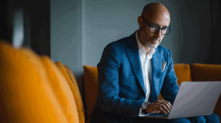 A man in a blue blazer and white shirt sits on a couch, focusing intently on his laptop. He wears round glasses and has a short beard. The background features soft, warm-colored cushions, creating a cozy atmosphere suitable for work or study.