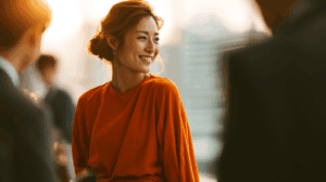 A woman wearing a bright orange dress smiles warmly as she engages in conversation. The background features blurred figures, suggesting a social gathering, with soft lighting that creates a warm atmosphere. Her hair is styled elegantly, adding to an overall sense of sophistication and joy.