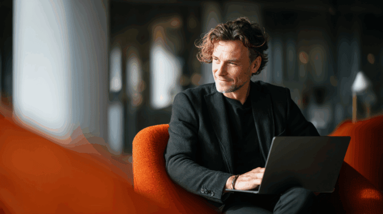 A man with curly hair sits comfortably in an orange armchair, working on a laptop. He wears a black blazer over a dark shirt and has a thoughtful expression, looking slightly to the side. The background features a modern interior with blurred elements, creating a professional atmosphere.