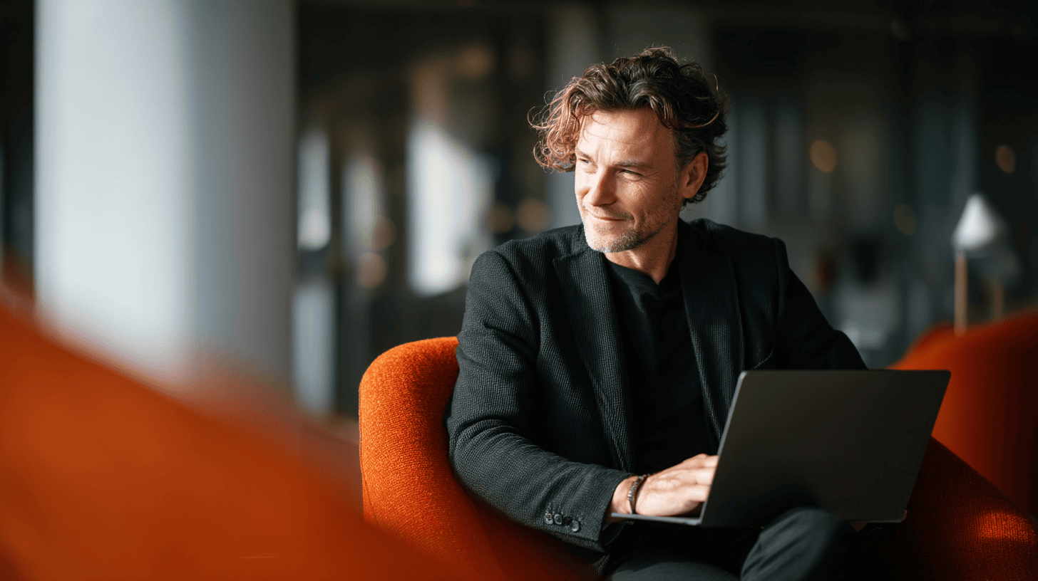 A man with curly hair sits comfortably in an orange armchair, working on a laptop. He wears a black blazer over a dark shirt and has a thoughtful expression, looking slightly to the side. The background features a modern interior with blurred elements, creating a professional atmosphere.