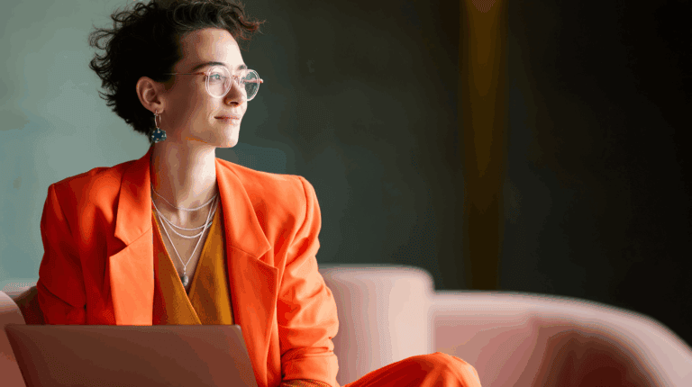 A professional woman with short, curly hair sits on a pink sofa, wearing an orange blazer and a brown top. She has stylish glasses and colorful earrings, gazing thoughtfully to the side while holding a laptop on her lap. The background features a soft-focus interior with muted colors.