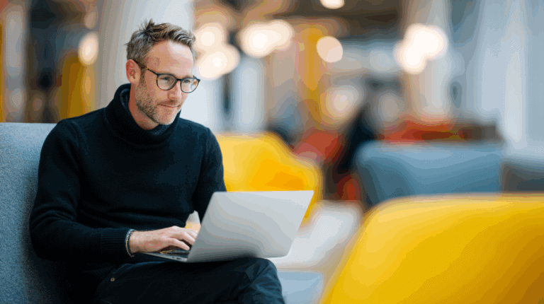 A man with short, tousled hair and glasses sits in a modern, comfortable setting, focused on his laptop. He wears a black turtleneck sweater and is surrounded by soft, colorful furniture. The background features soft lighting and a blurred view of other people engaged in conversation.