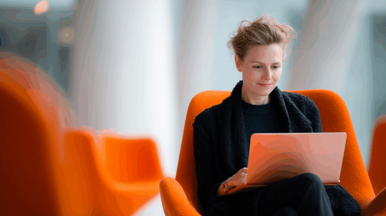 A woman sits comfortably in an orange chair, focused on her laptop. She has wavy hair and wears a black sweater with a shawl. The background features soft, blurred colors and shapes, suggesting a modern indoor space. The image conveys a sense of concentration and modern work environment.