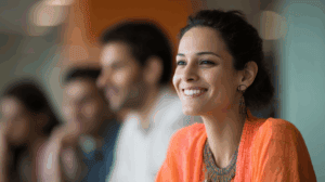 A woman smiling brightly, wearing an orange top and colorful jewelry, sits in a group discussion. Her joyful expression contrasts with the blurred figures of two people in the background, suggesting a collaborative or engaging environment. Soft lighting enhances the warm atmosphere.