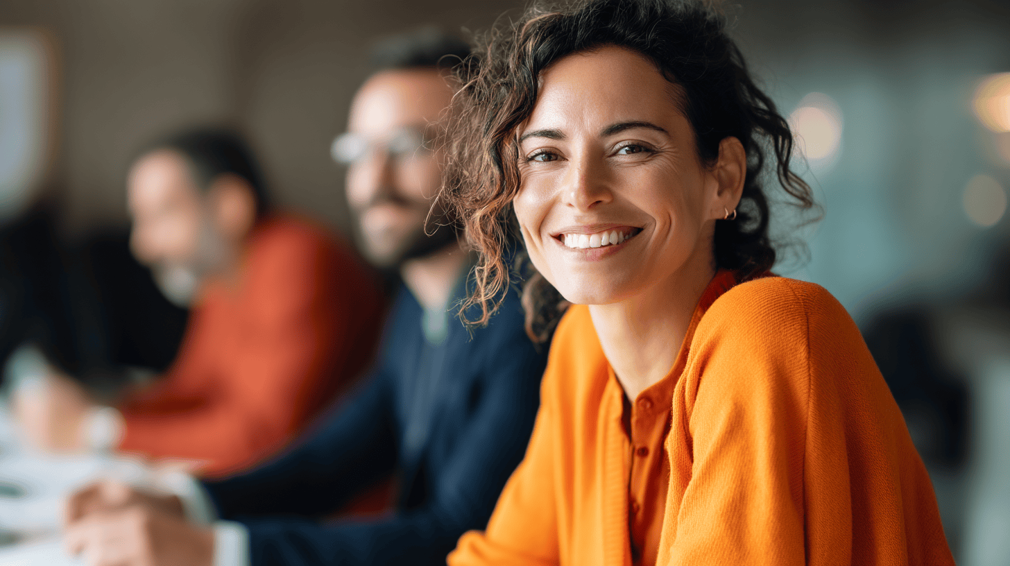 A smiling woman with curly hair wearing an orange sweater is seated at a table, engaged in conversation. Two blurred figures in the background are also present, suggesting a collaborative environment. The setting appears to be a modern, professional space.